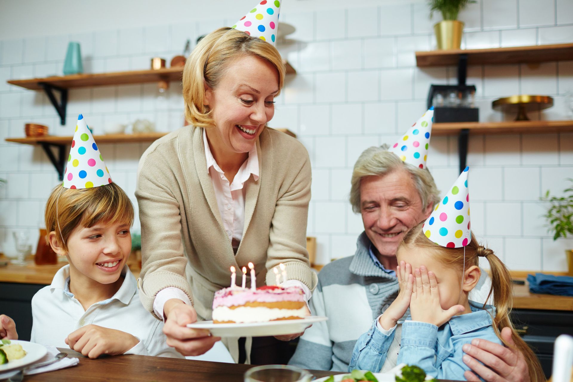Family celebrating a birthday in a kitchen: woman holding cake, child covering eyes, others in party hats.