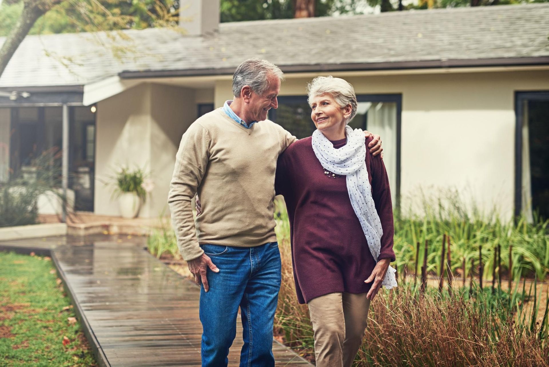 Senior couple walking together outside, man with arm around woman, smiling, near their house.