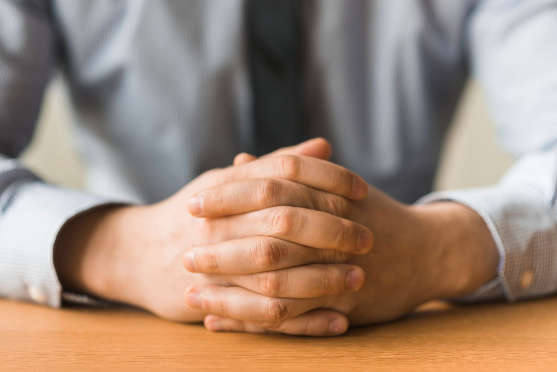 Person's clasped hands on a wooden table; a light blue shirt visible.
