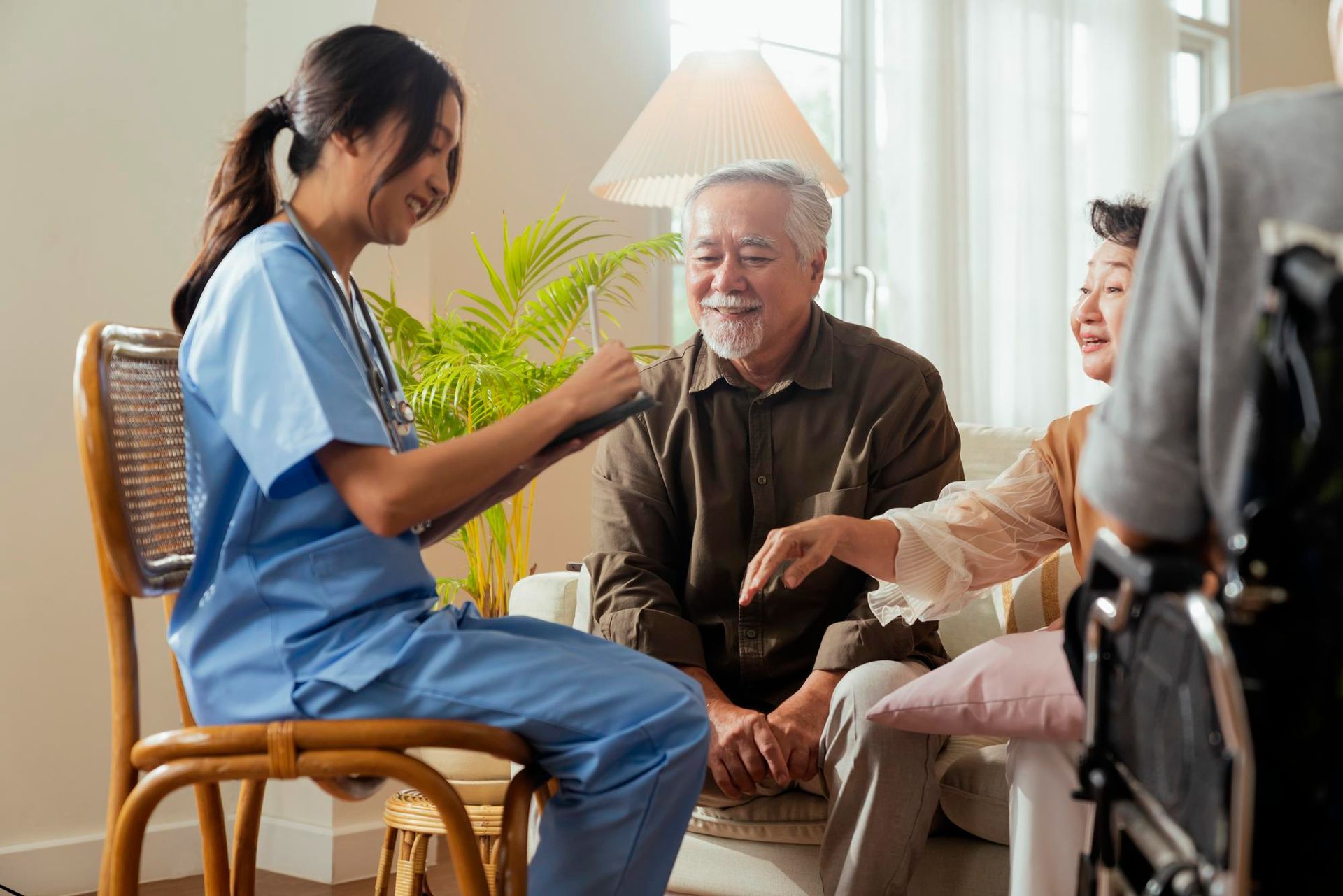 Nurse in scrubs examining elderly couple in a bright room. The woman in the wheelchair, smiling, touches the man's hand.