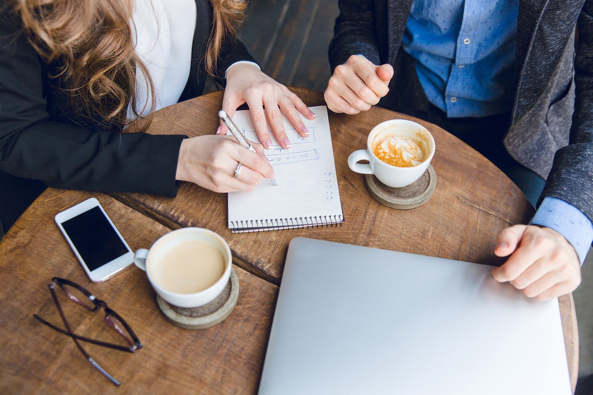 Two people at a table, one writing in a notebook, coffee, laptop, and phone.