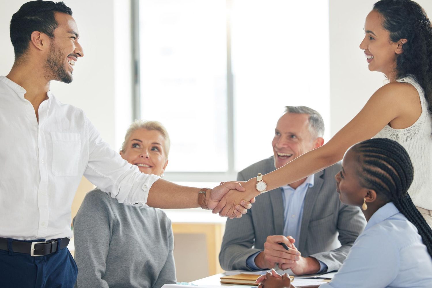 People in a business meeting, two shaking hands. Diverse group smiles in a bright room.