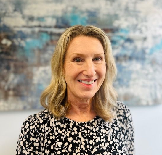 Woman in a black suit and pearl necklace smiles in a hallway.