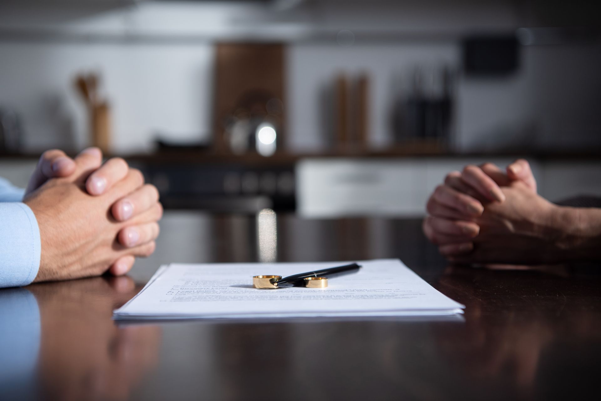Two people with hands clasped, facing a document with a pen and wedding rings on a table.