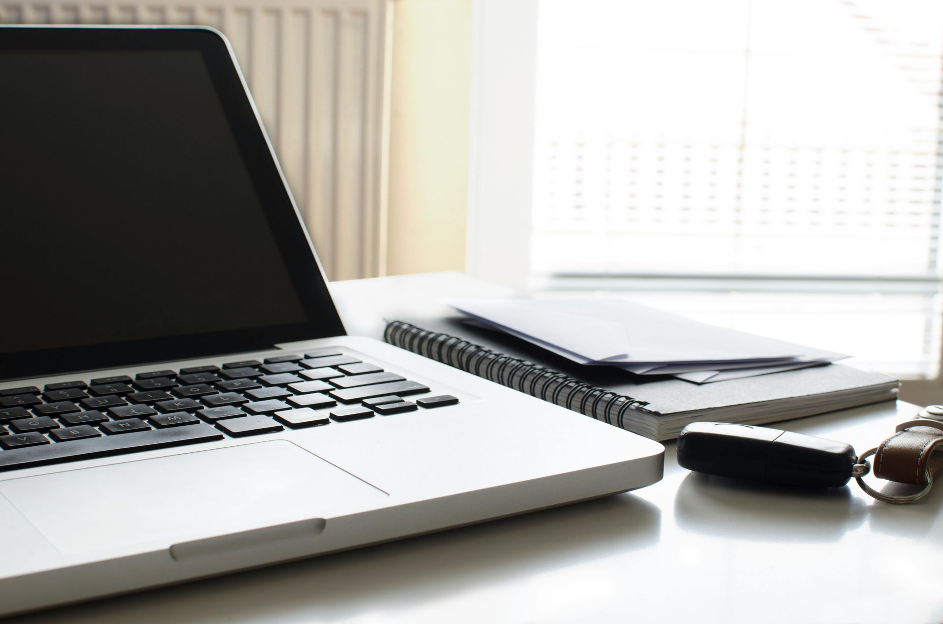 Laptop on desk with notepad, documents, and car keys near a window.