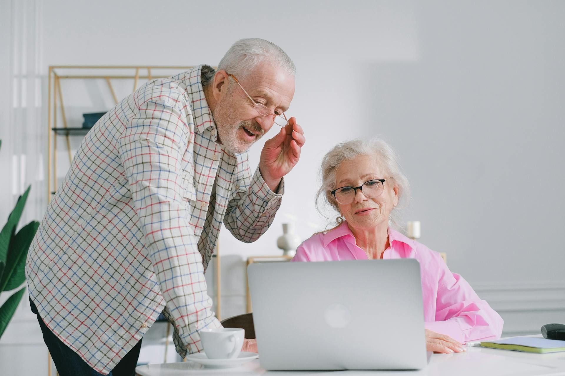 Elderly couple looking at laptop, man leaning in, wearing glasses and checkered shirt. Woman in pink, seated.