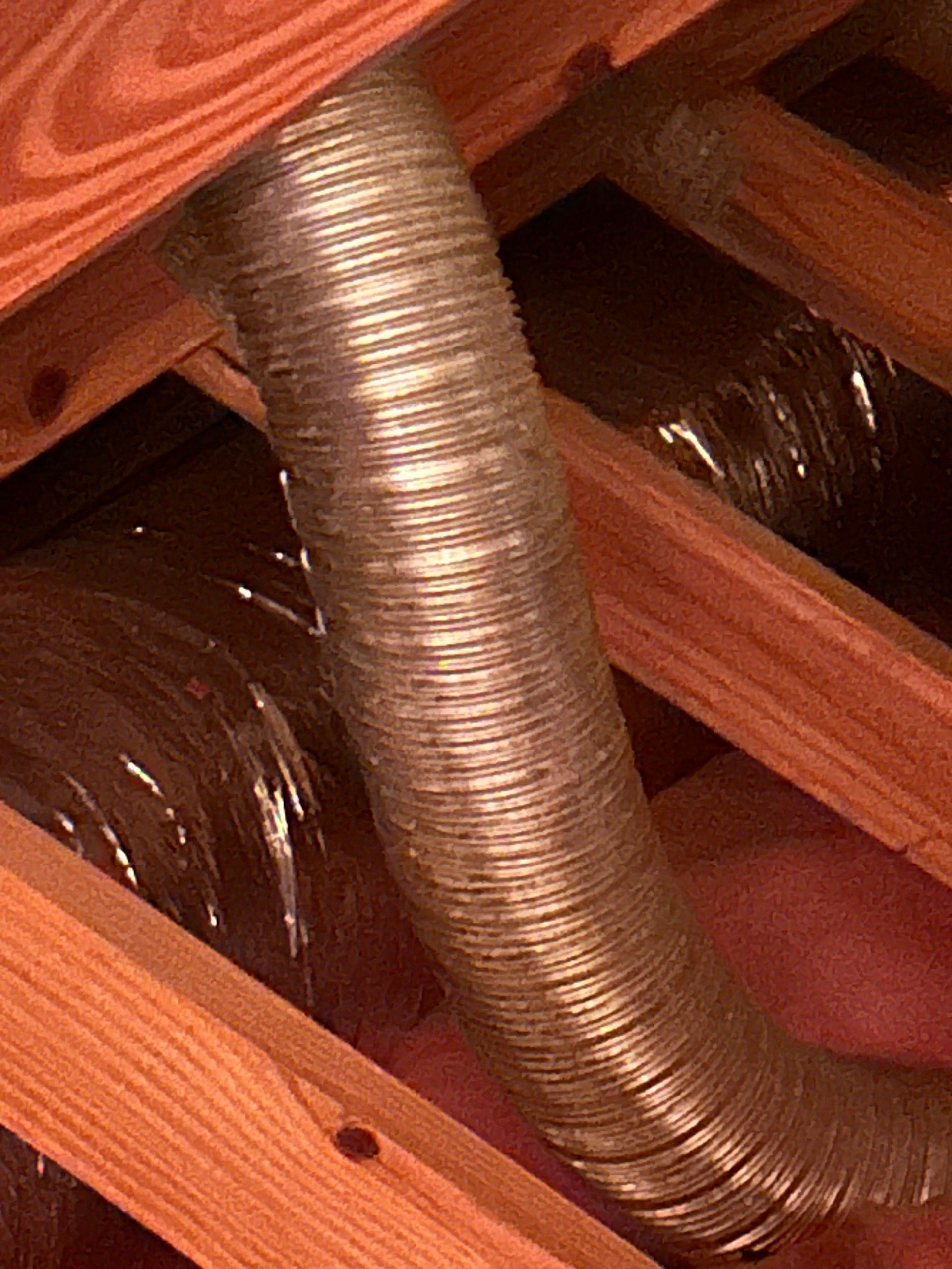 Flexible, silver-colored ventilation ductwork running through a wooden attic frame with pink insulation.