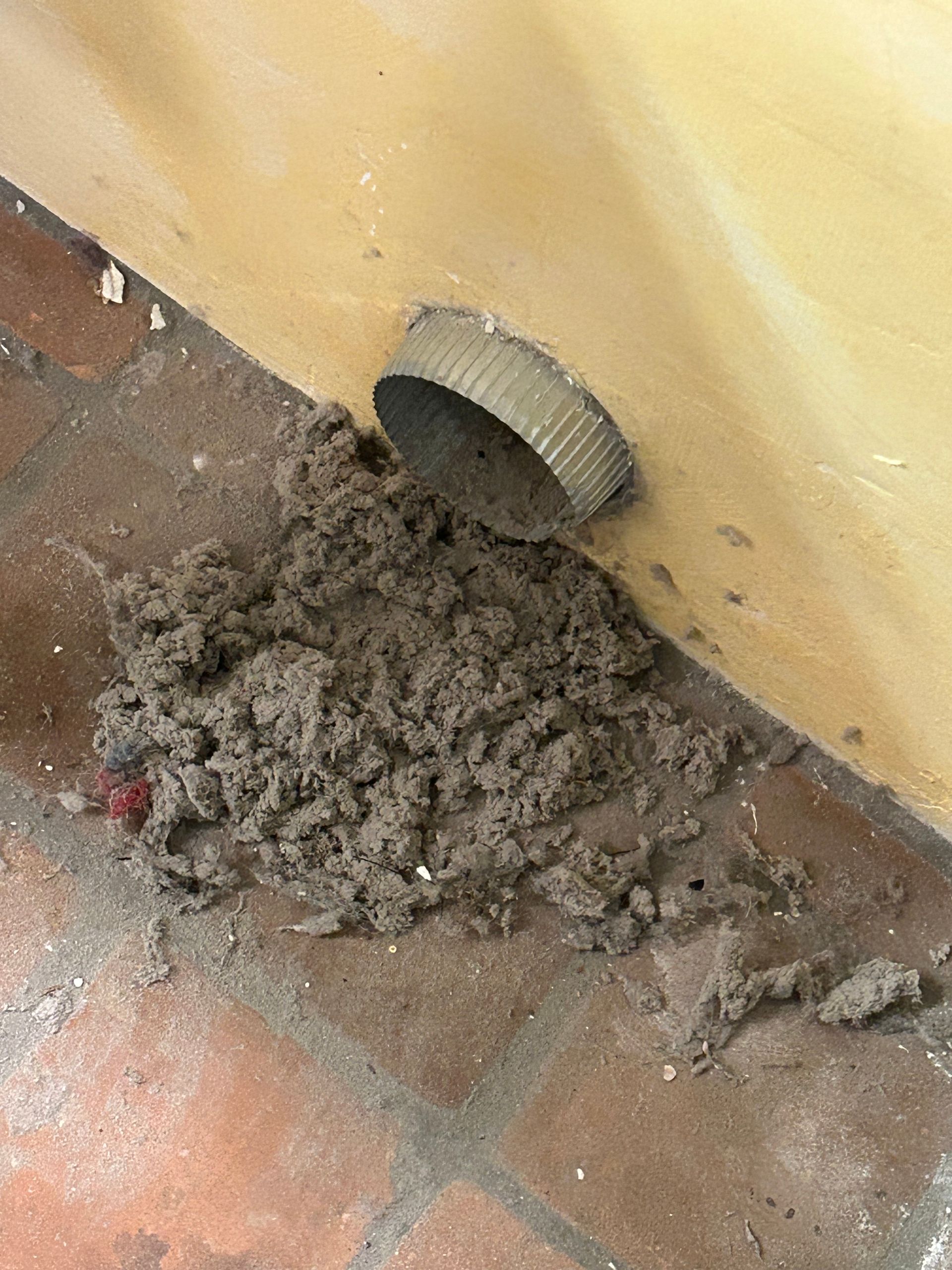 A large pile of gray dryer lint has accumulated on a tiled floor directly beneath a round metal dryer vent.
