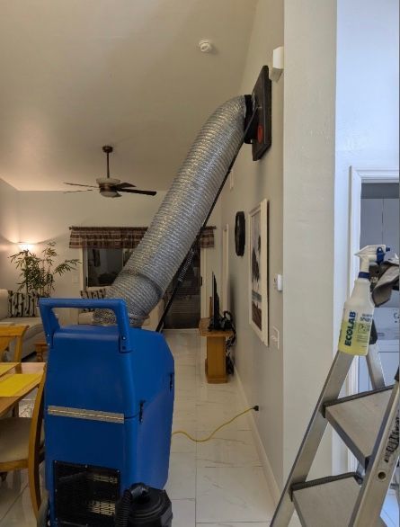 A blue portable industrial air scrubber is connected by a silver duct to an air vent mounted high on a white interior wall.
