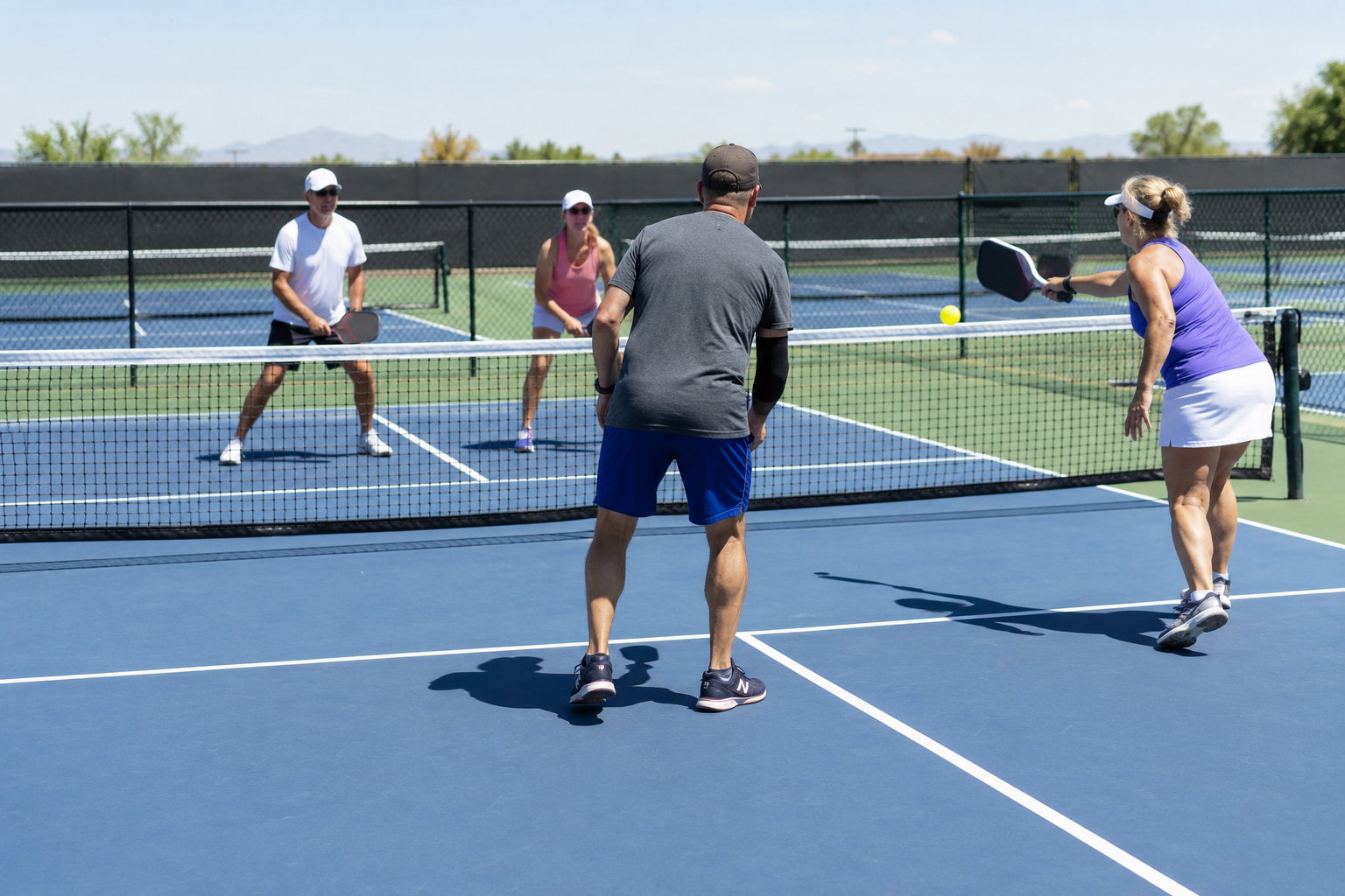 Four people play pickleball on a blue outdoor court, two near the net and two closer to the camera.