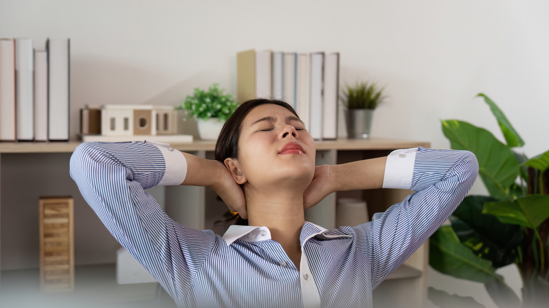 Woman relaxing with hands behind her head, eyes closed. Office setting with bookshelves and plants.