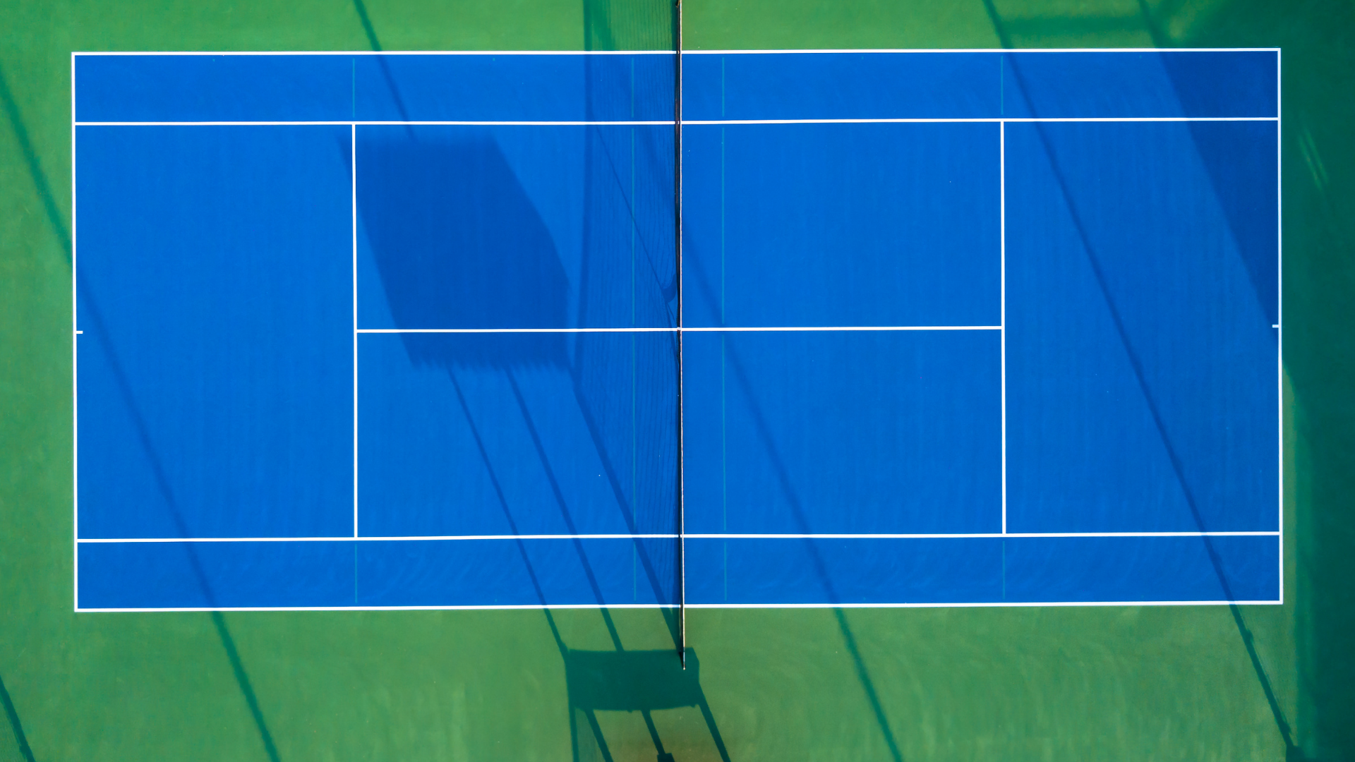 Blue tennis court, viewed from above, with net and white lines on green surrounding area.