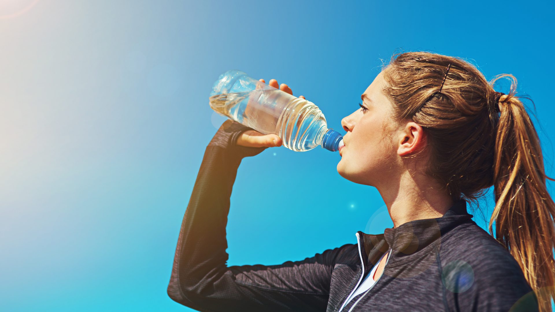 Woman drinks water from a bottle outdoors with a blue sky background.