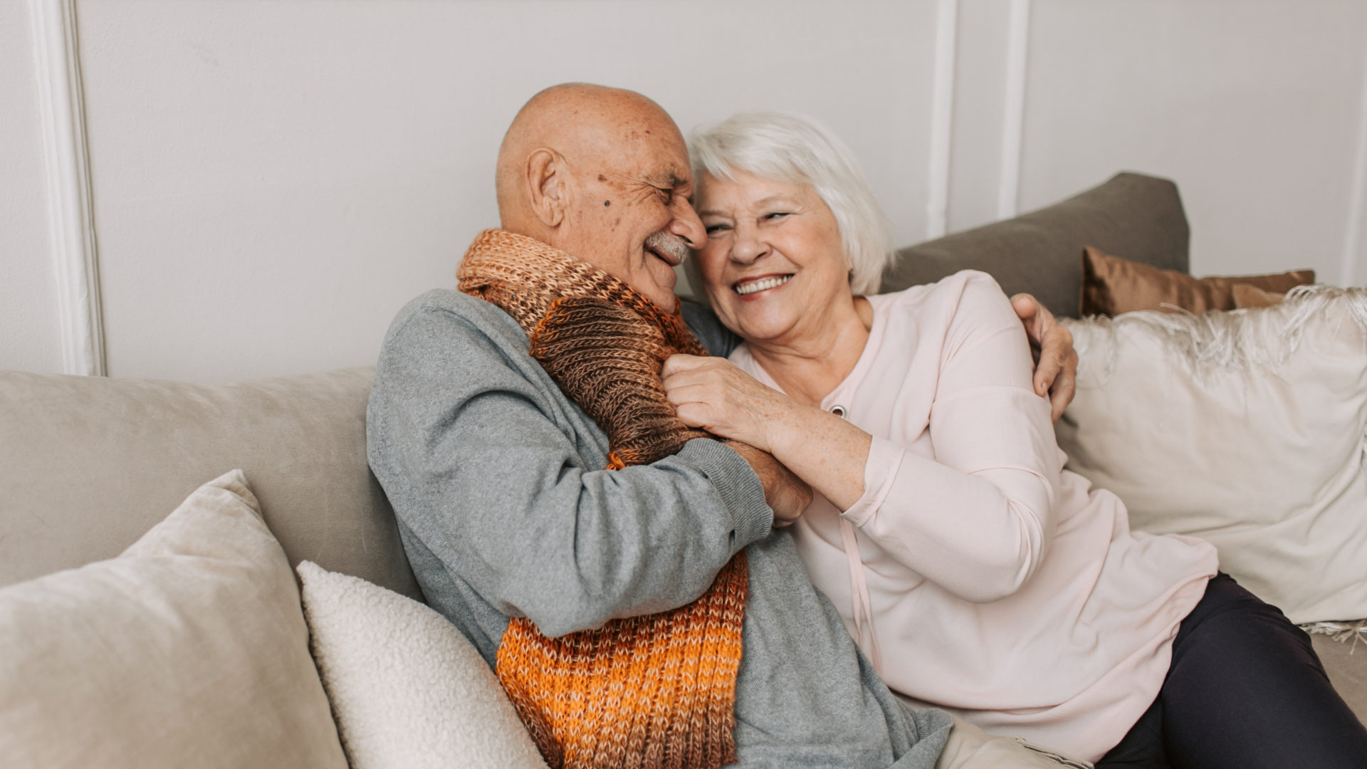 Elderly couple embracing and laughing on a couch. Man in scarf, woman in light-colored shirt.