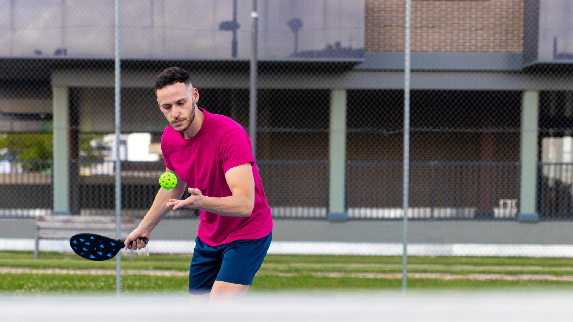 A person in a magenta shirt and blue shorts playing pickleball on an outdoor court, preparing to strike the ball.