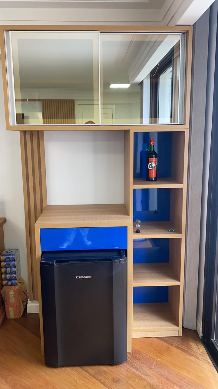 Uma pequena geladeira preta está ao lado de uma prateleira de madeira em uma sala.