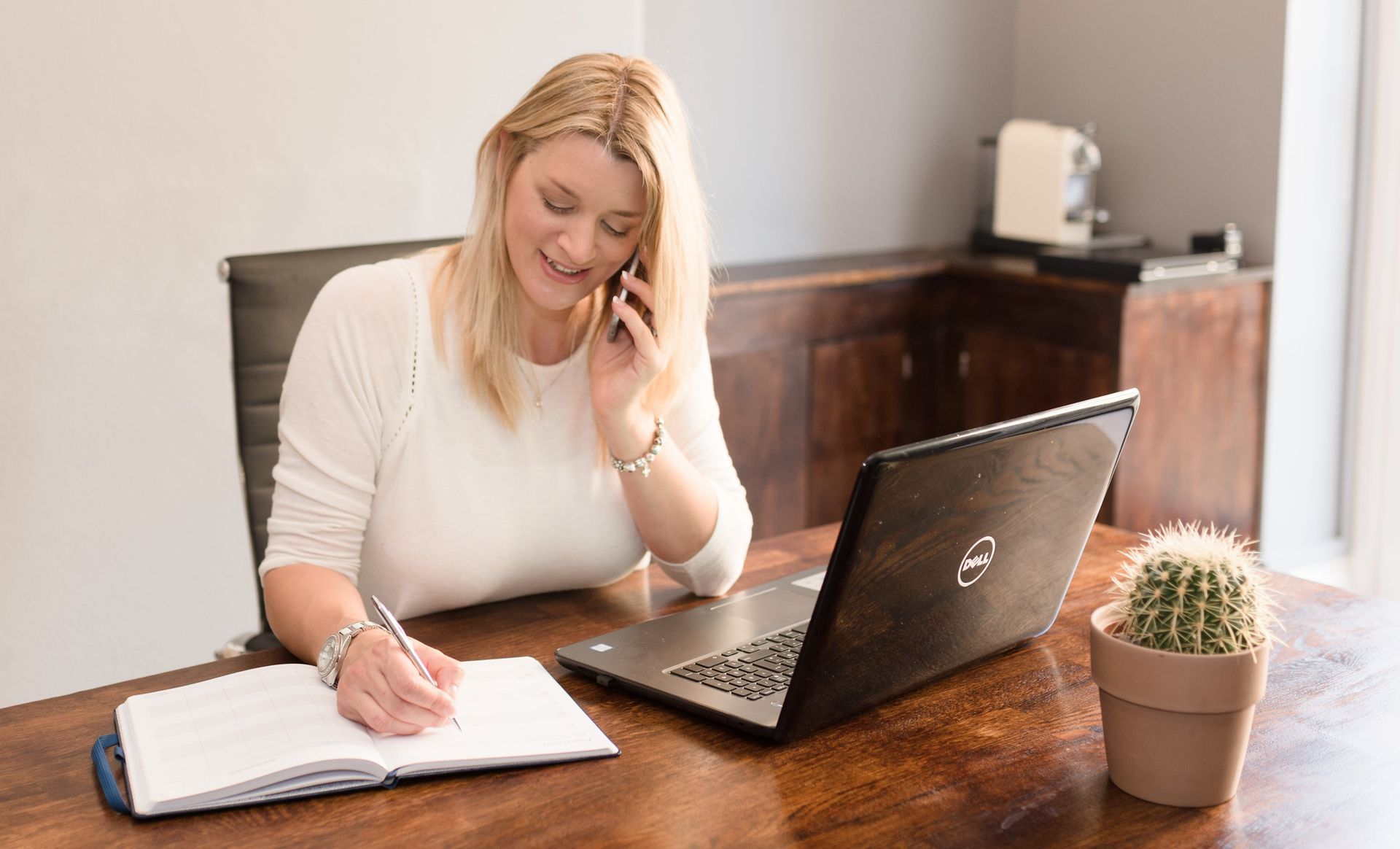 Kelly Owen sitting at a desk with an open laptop. She is talking on a mobile phone and making a note in a diary