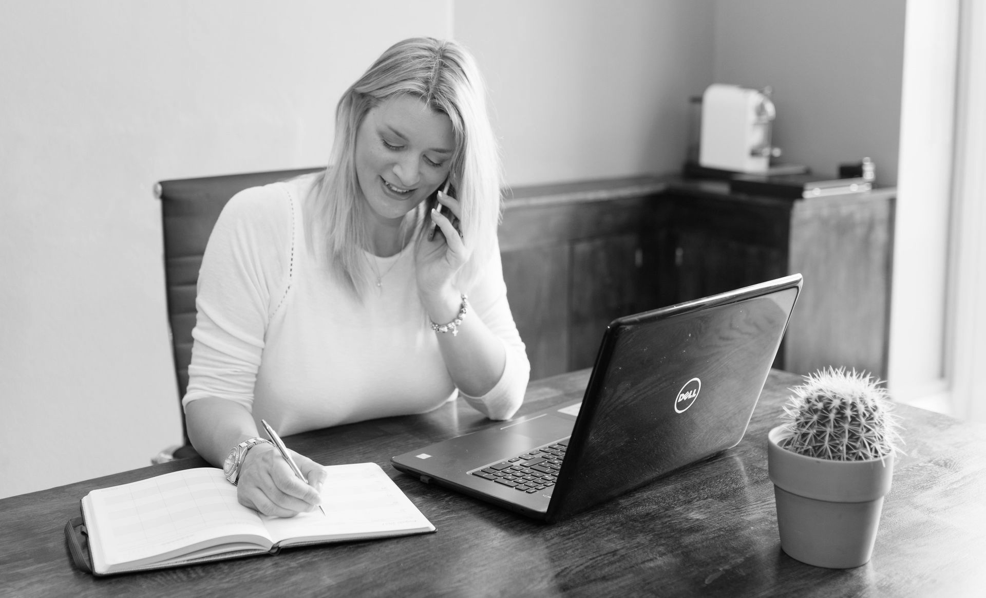 Kelly Owen sitting at a desk with an open laptop. She is talking on a mobile phone and making a note in a diary