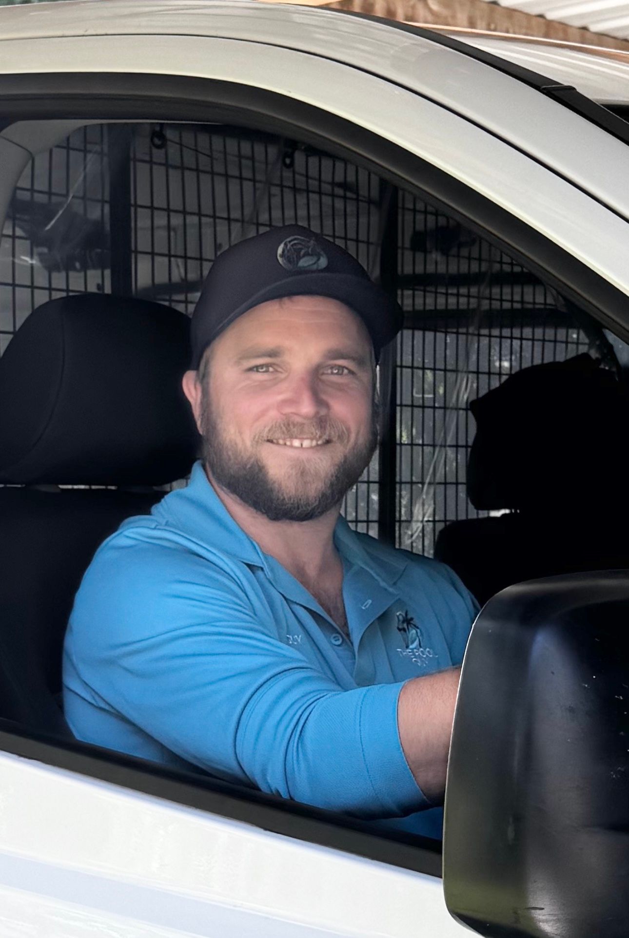 A person with a beard and baseball cap sits smiling in the driver’s seat of a white vehicle wearing a blue polo shirt. Pool Maintenance in Pacific Heights, QLD