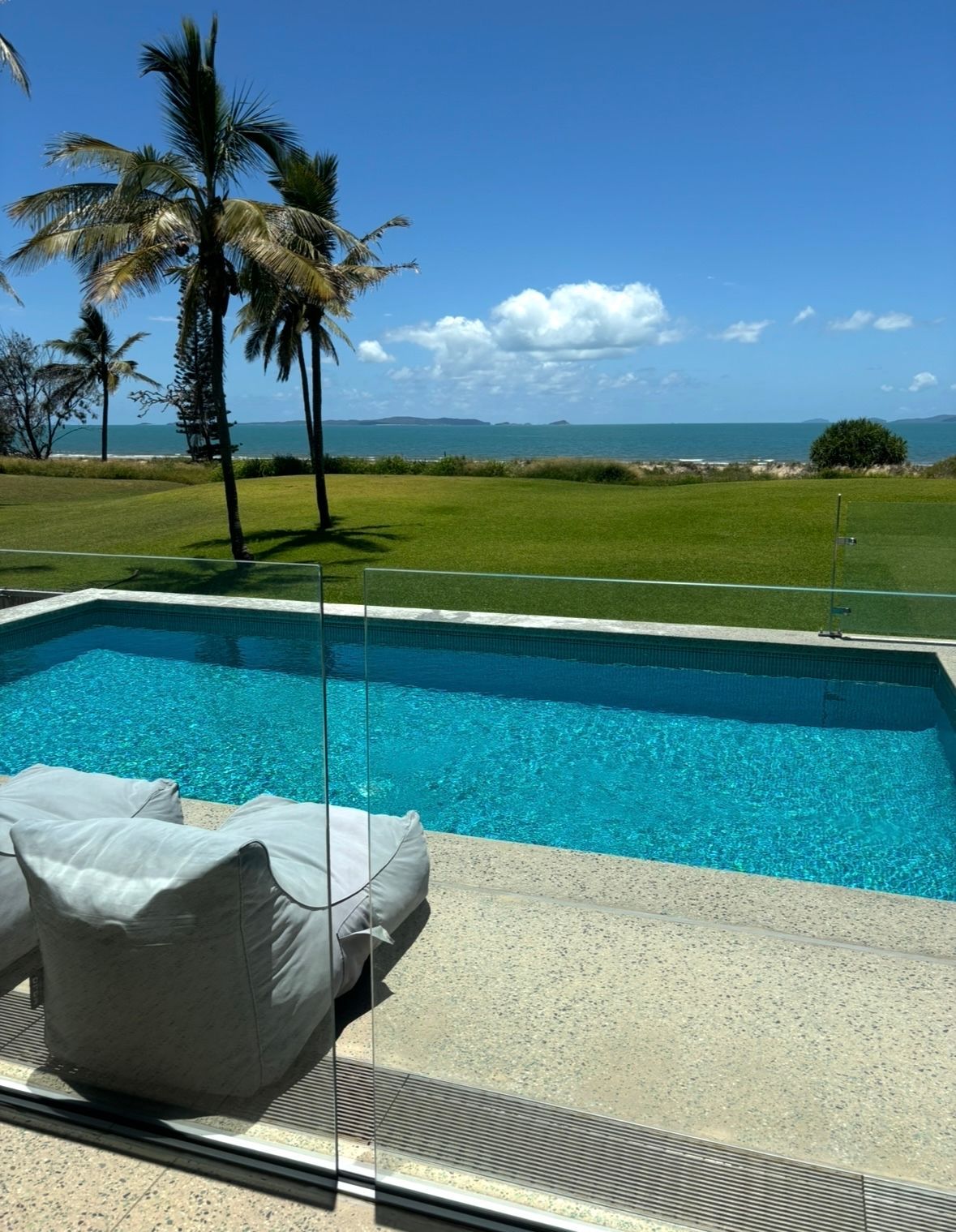 A swimming pool with turquoise water overlooks a green lawn, palm trees, and the ocean under a clear blue sky. Pool Maintenance in Pacific Heights, QLD
