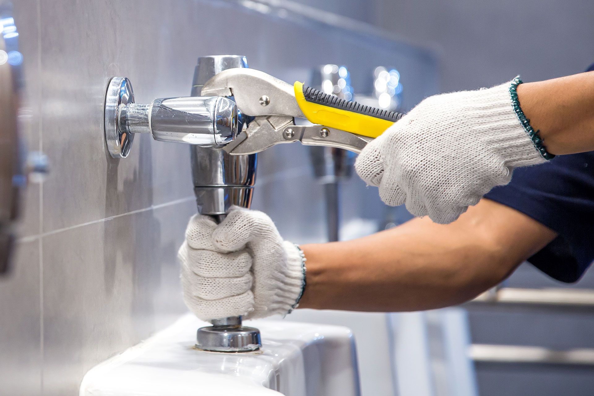 Plumber in gloves using pliers to tighten fixture on a urinal in a restroom.