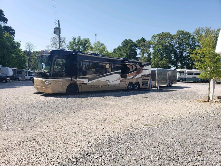 Large RV parked on gravel lot, beige and brown colors, trees in background.
