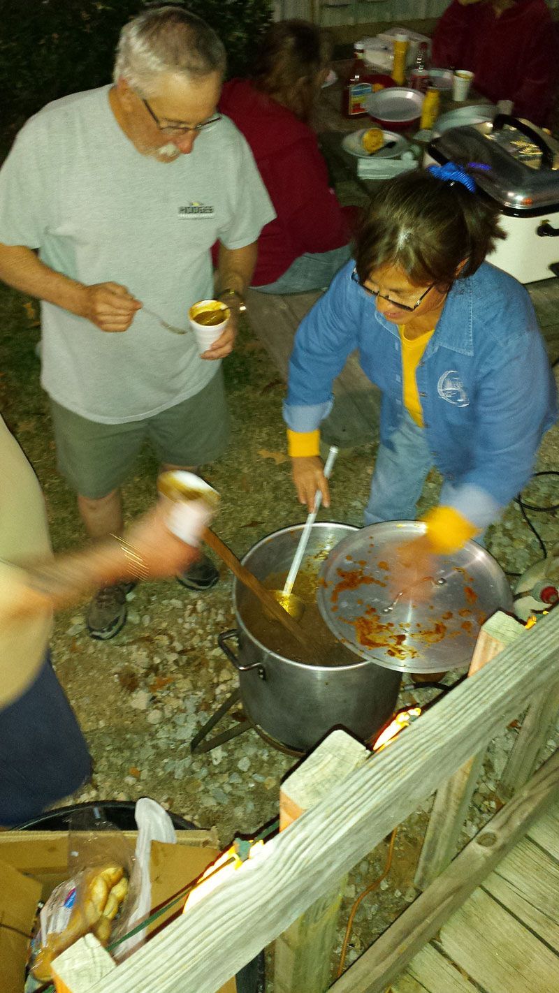 People serving food from a large pot outdoors. Woman in blue jacket stirs, man eats from a cup.