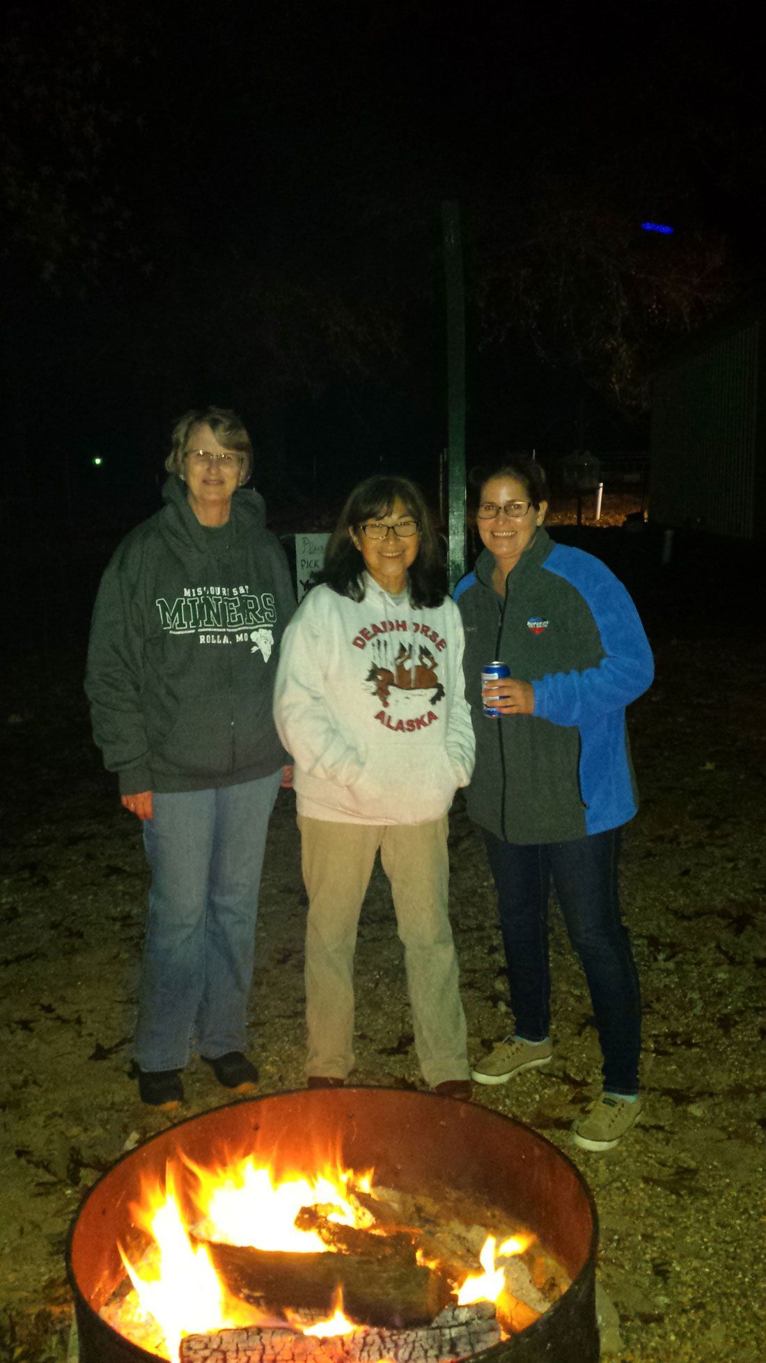 Three women stand by a fire pit at night. They wear casual clothes; the flames are bright.