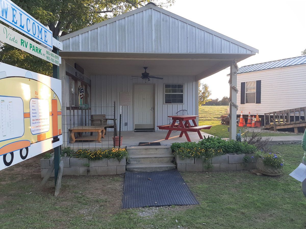 Small white cabin with porch and picnic table, RV Park sign on the left, sunny day.
