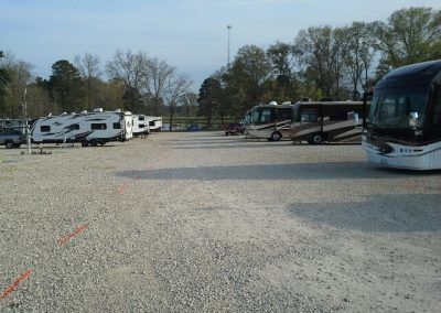 RV and bus parking lot, gravel surface. Vehicles parked; a few trees in the background. Clear sky.