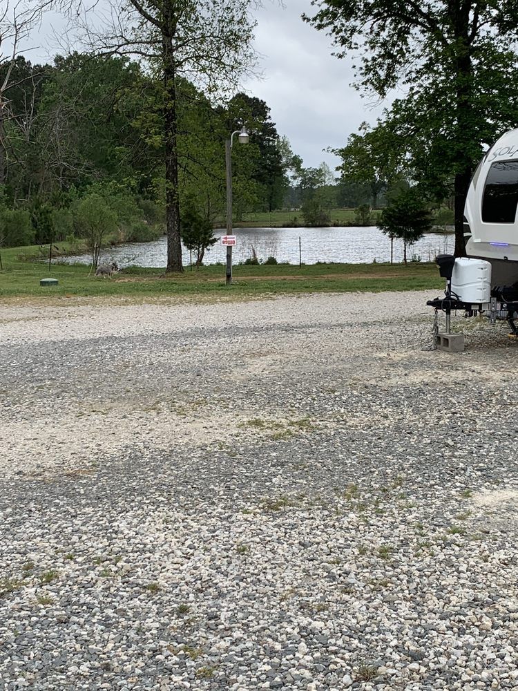 A gravel campsite with a camper, overlooking a pond and trees under a cloudy sky.
