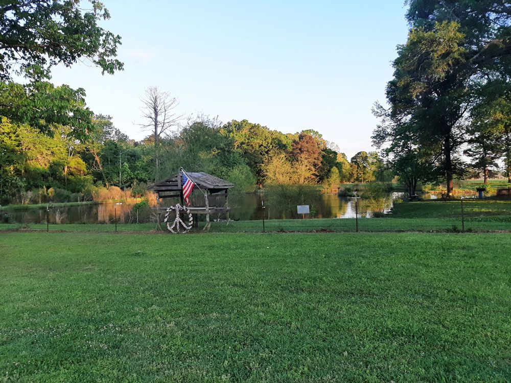 Lawn with a gazebo and American flag near a pond, surrounded by trees.