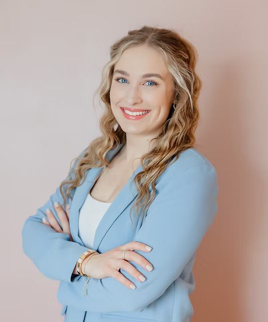Blonde woman in blue blazer with arms crossed, smiling. Pink background.