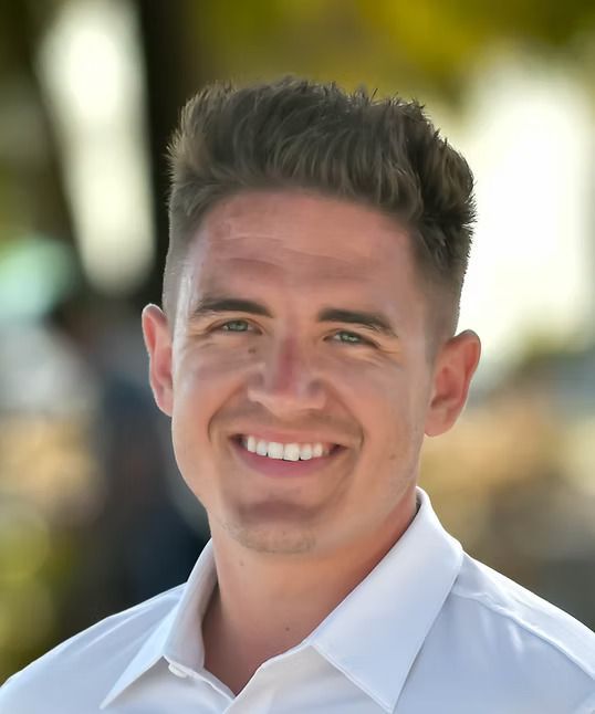 Man with short, brown hair, smiling, wearing a white shirt, outdoors with a blurred background.