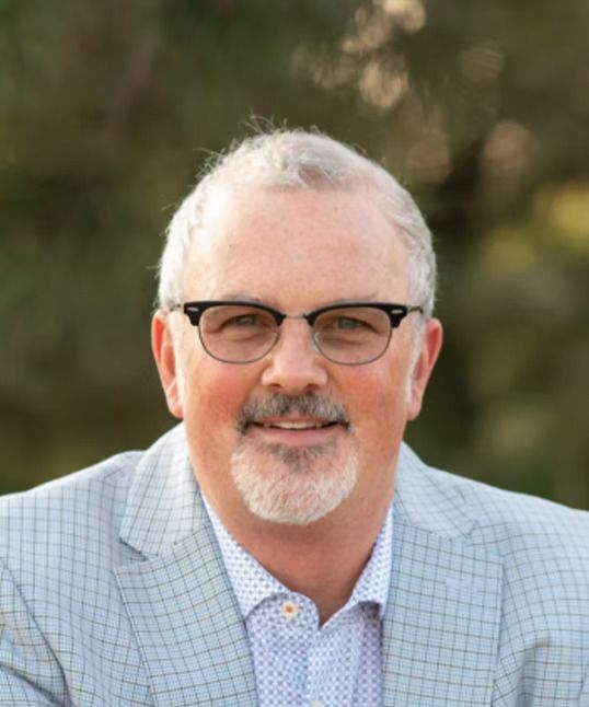 Man with graying hair and glasses, wearing a light blue blazer, smiling outdoors.