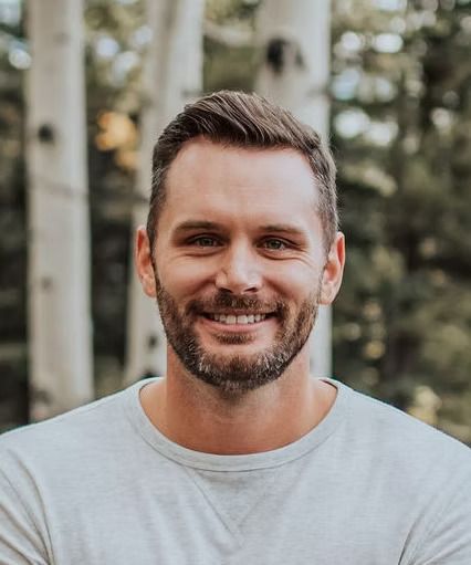Smiling man with short brown hair and a beard, wearing a light gray shirt, in front of a blurred forest backdrop.