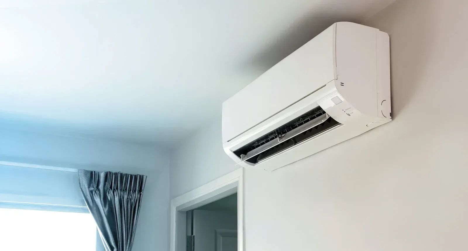 White wall-mounted air conditioning unit in a room with window and open doorway.
