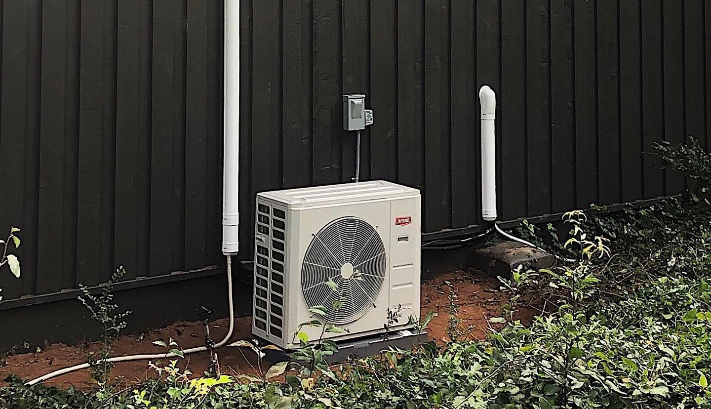 A white air conditioner is sitting on the side of a building next to a fence.