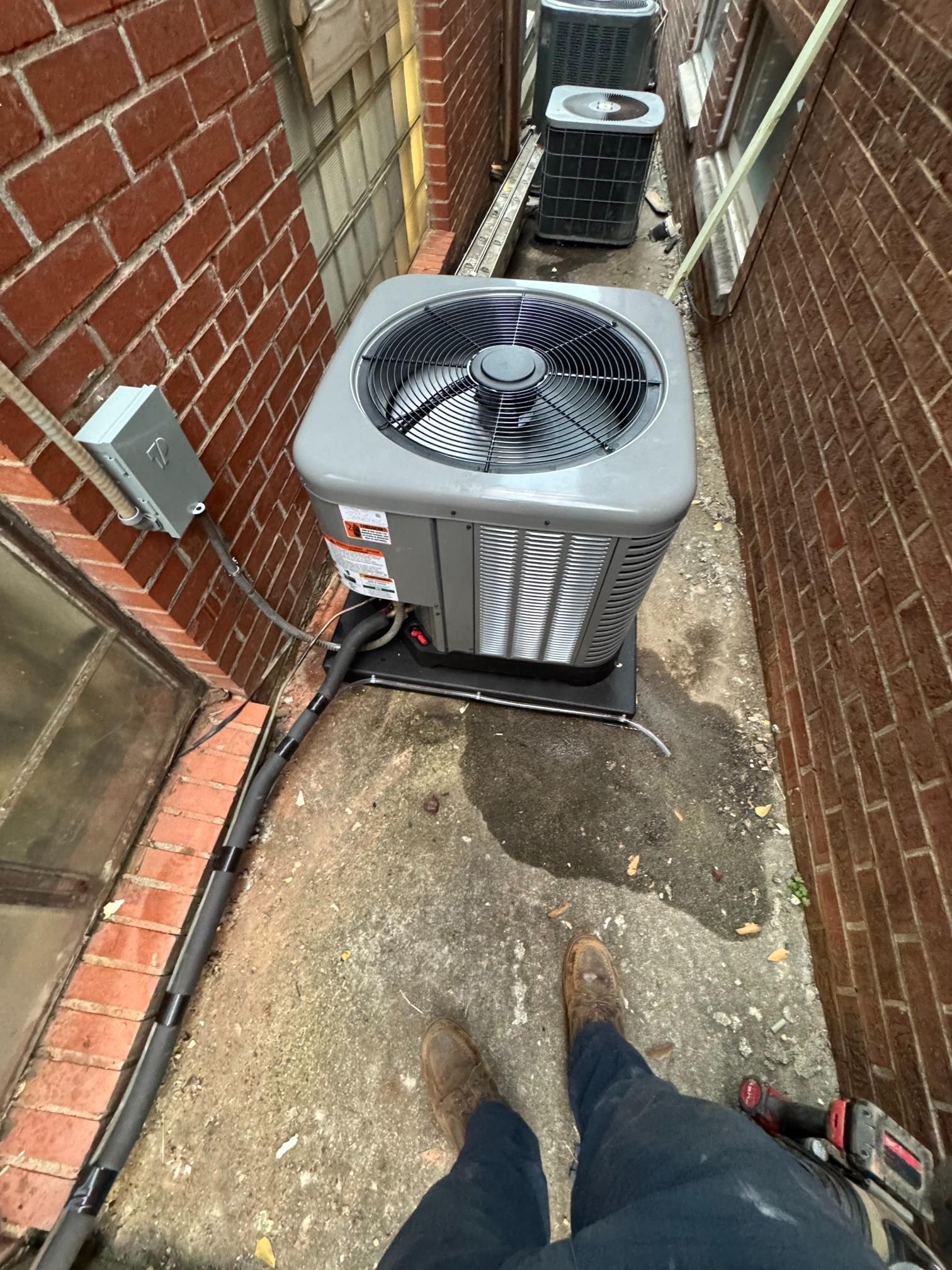 Outdoor HVAC units in a narrow alley beside a brick wall, with a person’s feet at the bottom edge.