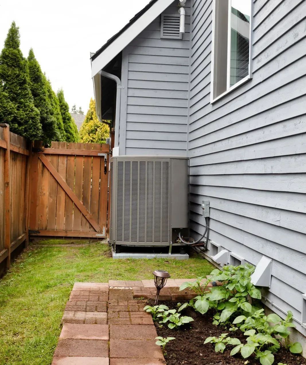 AC unit next to gray house, small yard with brick path and wooden fence.