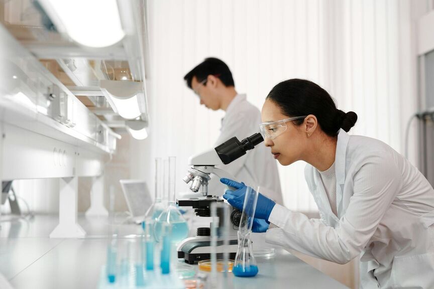 Two lab technicians in a lab with microscopes and test tubes; one looking through microscope, one working.