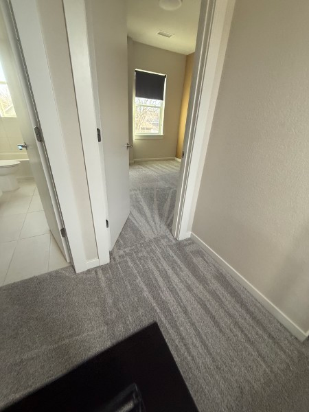 Hallway with beige walls, gray carpet, and a window at the end beside a bathroom door
