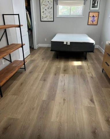 Empty bedroom with wood flooring, gray walls, a bed, window, wall art, and a small dresser.