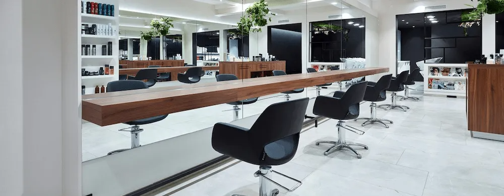 L'interno di un salone da parrucchiere con lunghi tavoli in legno, sedie nere e pareti bianche.