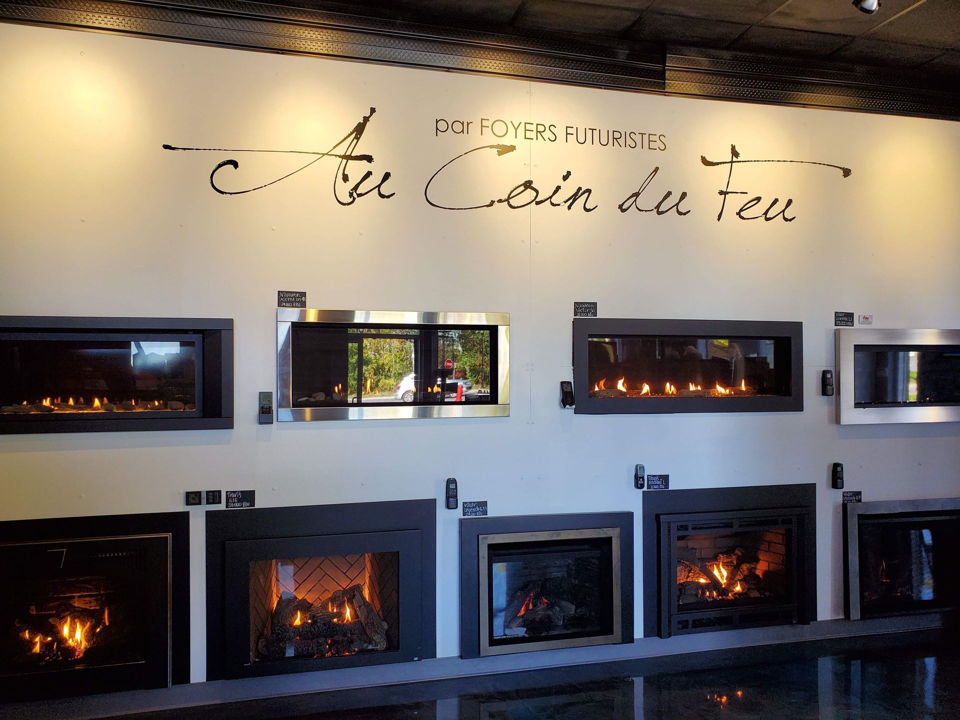 Cheminées exposées dans une salle d'exposition, flammes illuminées sur un mur blanc. Inscription « Au Coin du Feu ».