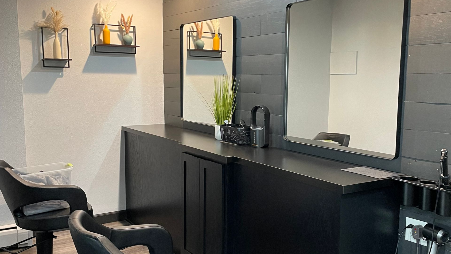 Hair salon station: black counter, mirrors, chairs, decorative shelves with product, neutral colors.