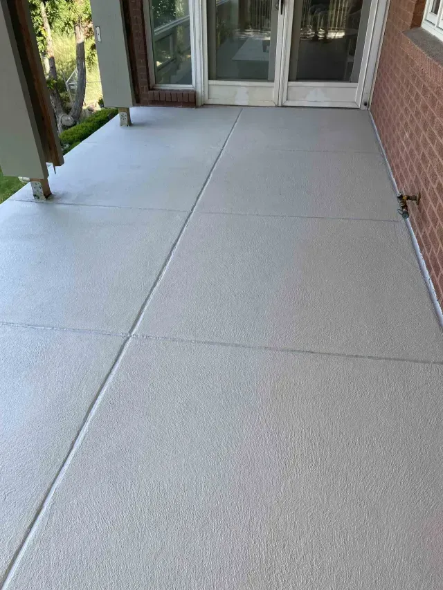A clean, light gray concrete patio surface with rectangular control joints, adjacent to a brick wall and a glass door.