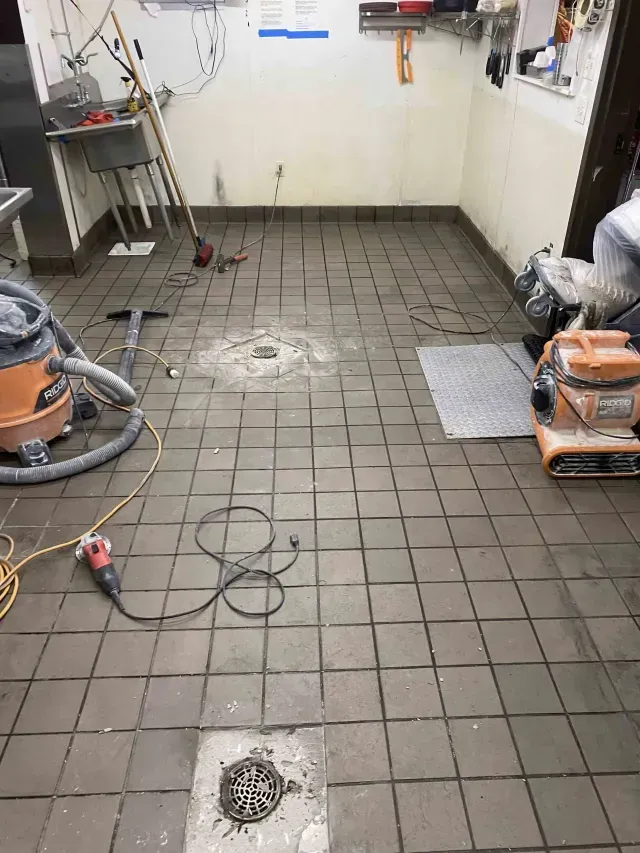A commercial kitchen floor undergoing repair with two floor drains, a vacuum, and power tools scattered about.