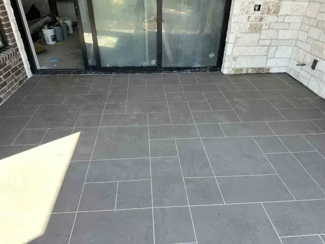 A gray stone patio featuring a rectangular paver pattern, adjacent to a brick wall and a glass sliding door.