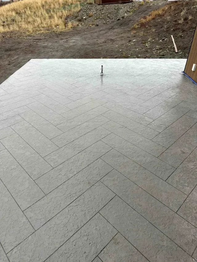 A gray concrete patio featuring a herringbone pattern, situated outdoors in front of an unfinished dirt yard.